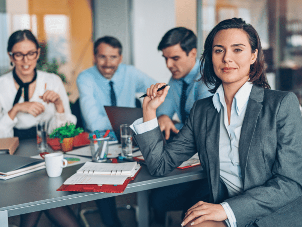 A confident businesswoman in a suit sits at a conference table with three colleagues in the background, all appearing engaged in a meeting. Papers, pens, and coffee cups are on the table.