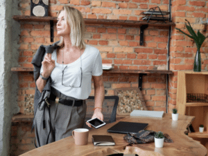 A woman with blond hair stands in a modern office with exposed brick walls, holding a smartphone and blazer, with a laptop, mug, and other items on a wooden desk. She looks thoughtfully into the distance.